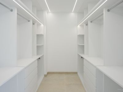 Ruime witte inloopkast met op maat gemaakte planken, laden en hangstangen, verlicht door geïntegreerde LED-strips en een lichte tegelvloer.
