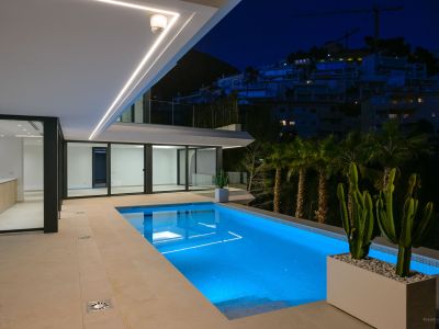 Verlicht zwembad 's nachts met levendig blauw onderwaterlicht en een ruim betegeld terras. Moderne lineaire plafondverlichting accentueert de architectuur, met uitzicht op een heuvel met verre gebouwen.