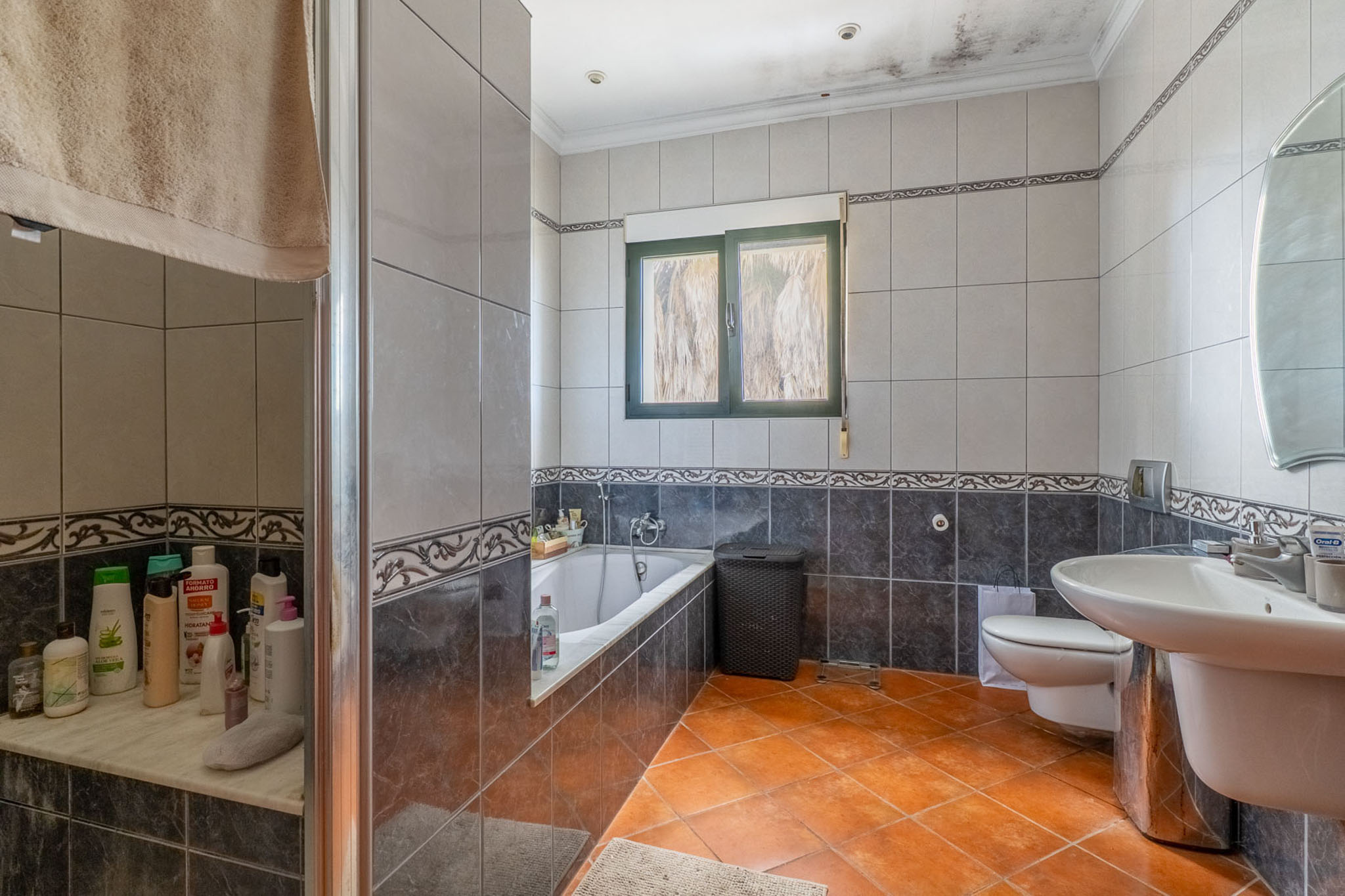 Complete badkamer met terracotta vloertegels, grijze wandtegels met sierranden, een ligbad, glazen douchecabine, wastafel en een groen kozijnraam.