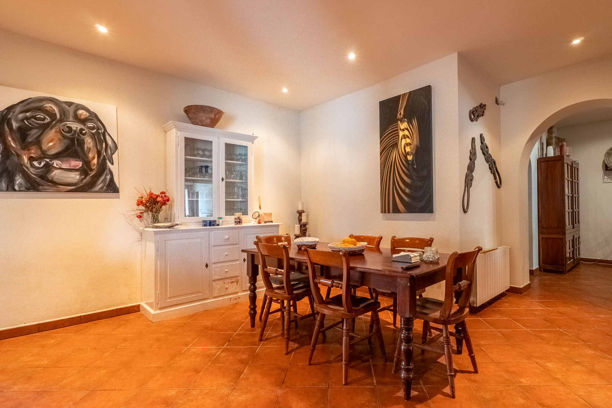 Eetkamer met donkere houten tafel en zes stoelen, terracotta tegelvloer en witte muren met inbouwspots. Een witte vitrinekast en twee decoratieve schilderijen sieren de ruimte.