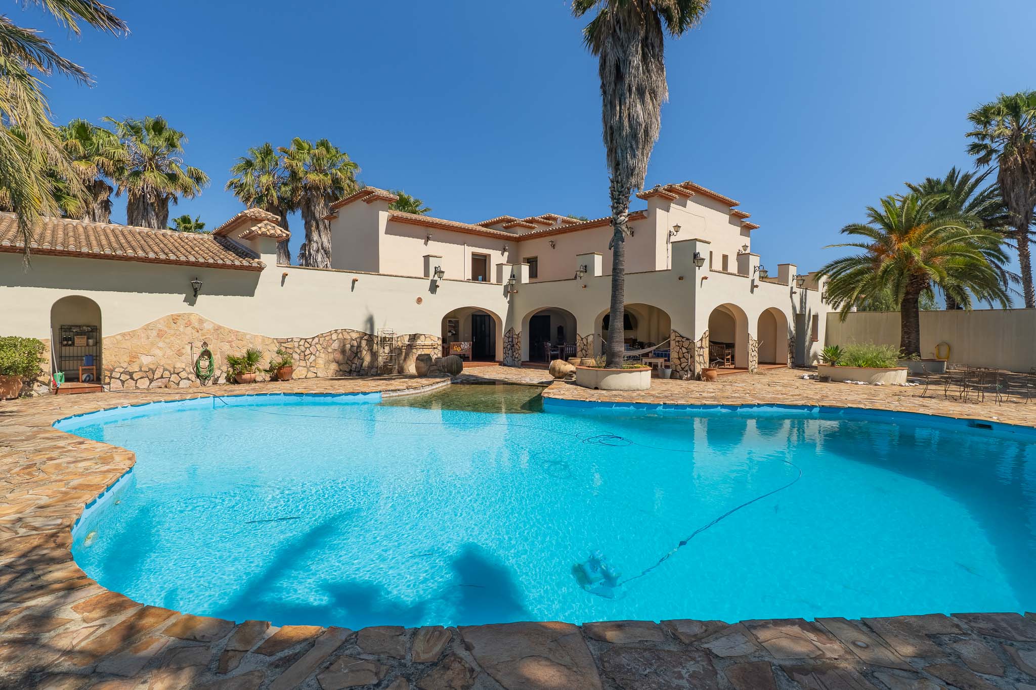 Exterieur van de mediterrane villa met een onregelmatig gevormd privézwembad, natuurstenen randen en omringd door palmbomen onder een helderblauwe lucht.