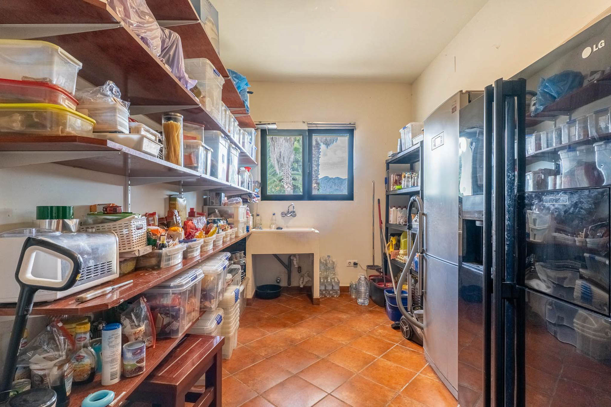 Functionele bijkeuken met uitgebreide schappen, een spoelbak en twee grote koelkasten. Terracotta tegelvloer en een raam met uitzicht op palmbomen en bergen.