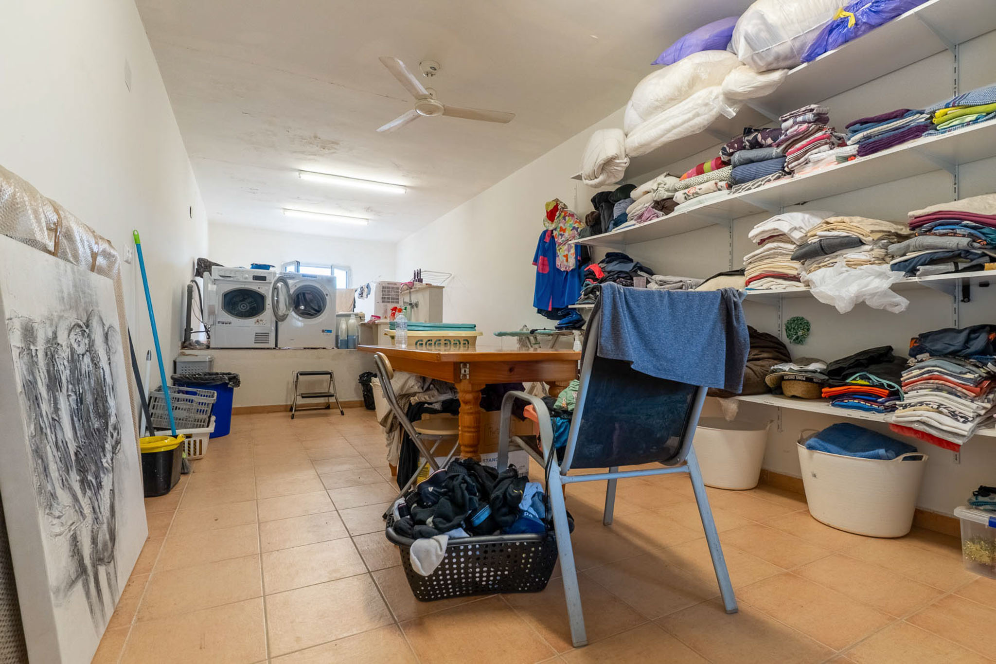Functionele wasruimte met twee wasmachines, droger en open planken voor wasgoed. Een centrale houten tafel biedt een praktische werkruimte, aangevuld met tegelvloer en plafondventilator.