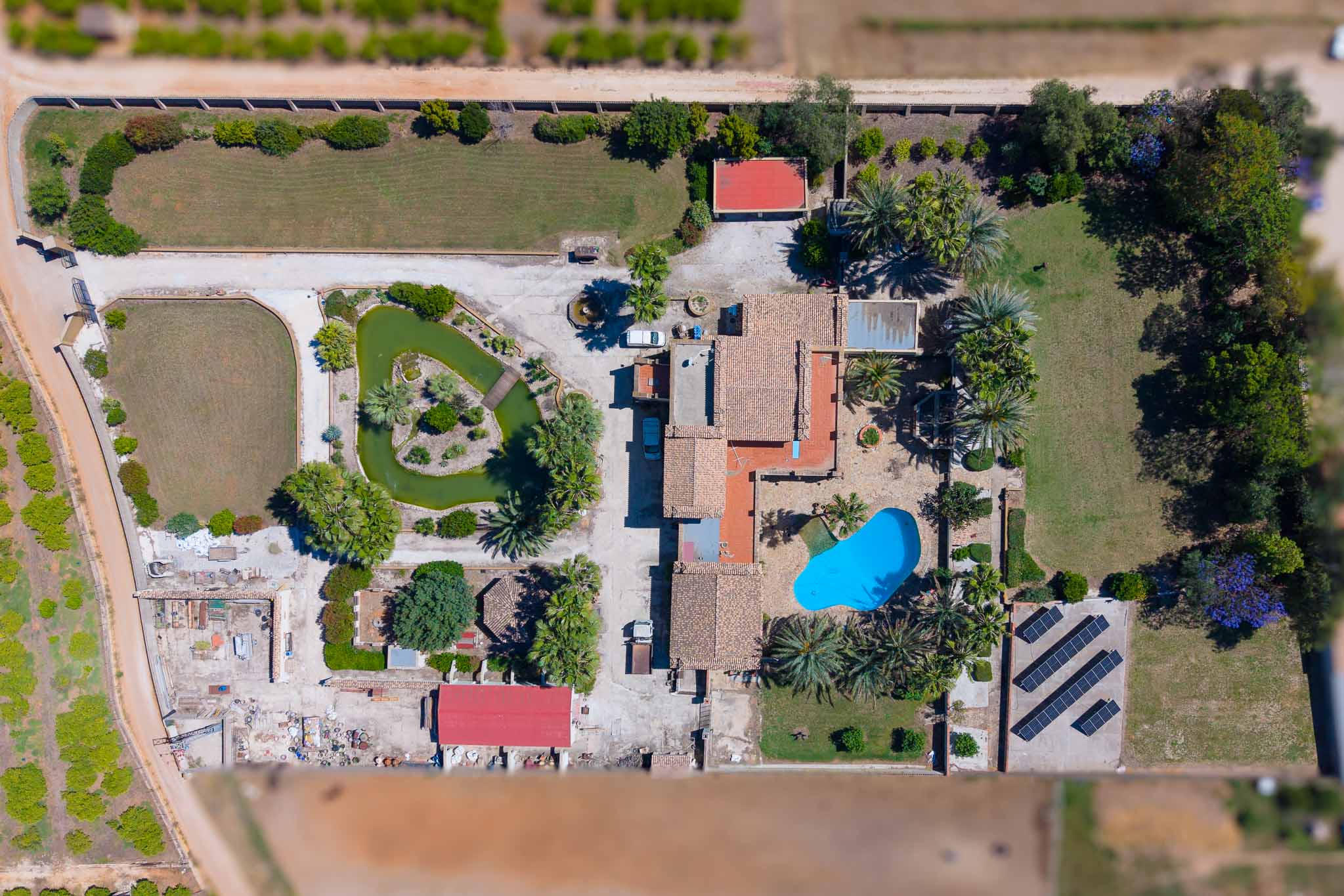 Gedetailleerde luchtfoto van het uitgestrekte perceel, met de hoofdvilla, blauw zwembad, vijver met eiland en de zonnepaneelinstallatie.