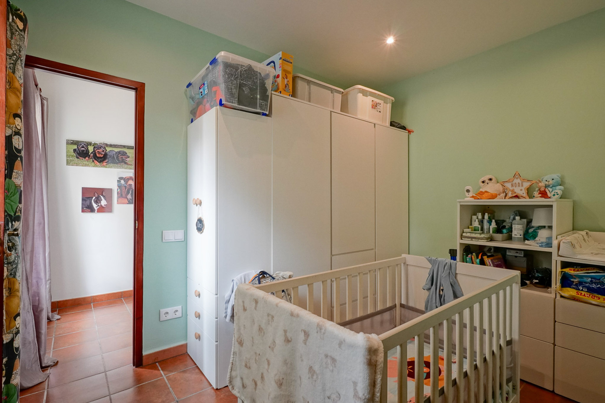 Kinderkamer met lichtgroene muren en terracotta tegelvloer. Voorzien van een wit ledikant, ruime witte kledingkasten en een open rek, verlicht door een inbouwspot in het plafond.