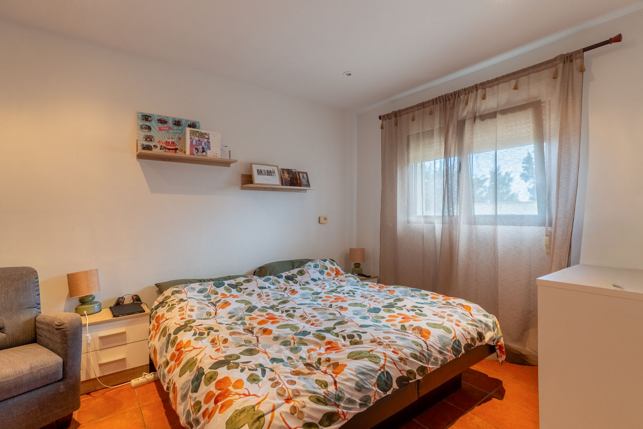 Lichte slaapkamer met tweepersoonsbed, kleurrijk bladpatroon dekbed en terracotta tegelvloer. Natuurlijk licht valt binnen via een raam met vitrage, aangevuld met een nachtkastje en wandplanken.