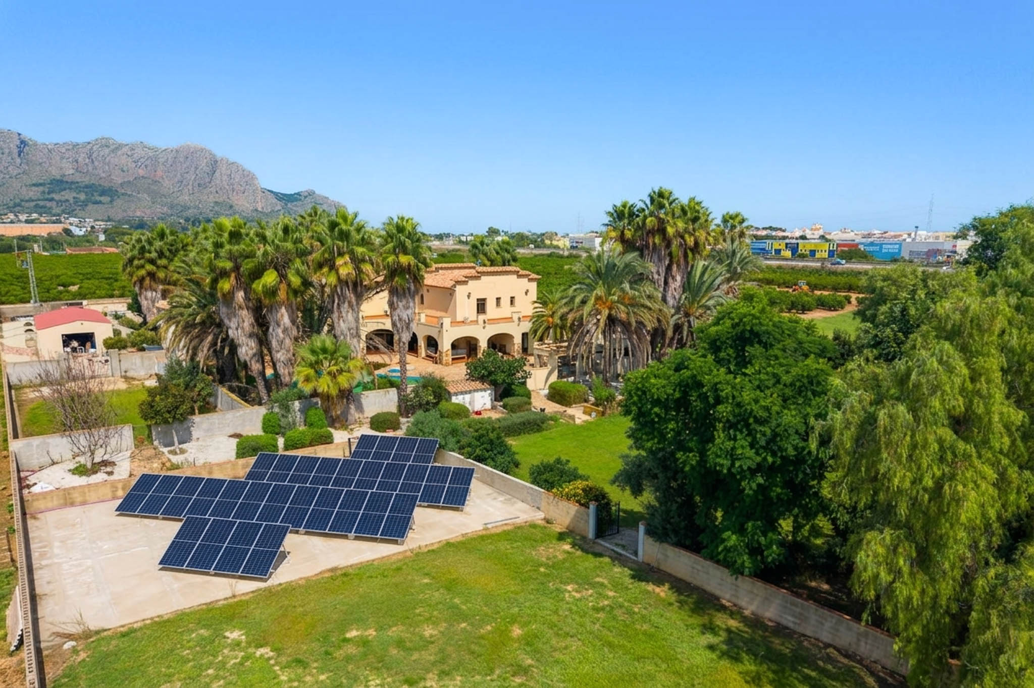 Luchtfoto van de traditionele villa, omringd door palmbomen en weelderige tuinen, met een opvallende zonnepaneleninstallatie en verre berguitzichten onder een heldere hemel.