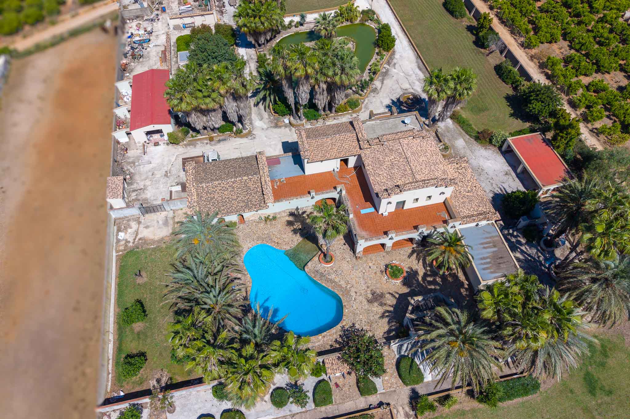 Luchtfoto van het landgoed met een onregelmatig blauw zwembad, omgeven door palmbomen en tuinen. Meerdere gebouwen met pannendaken en een hartvormige vijver zijn zichtbaar.