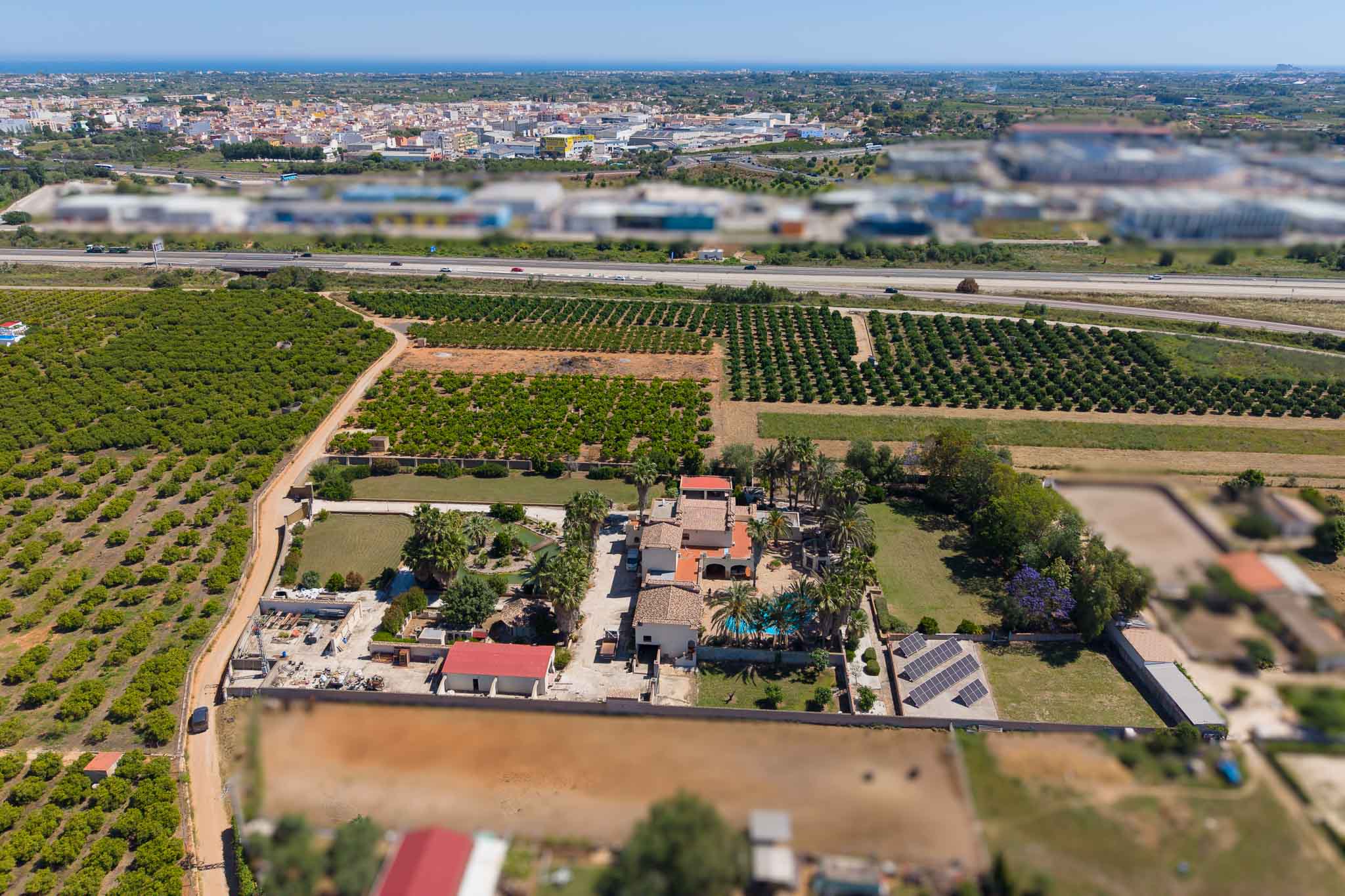Panoramische luchtfoto van het pand, met privézwembad, zonnepanelen en uitgestrekte sinaasappelboomgaarden. Snelweg, stad en de verre Middellandse Zee completeren het landschap.