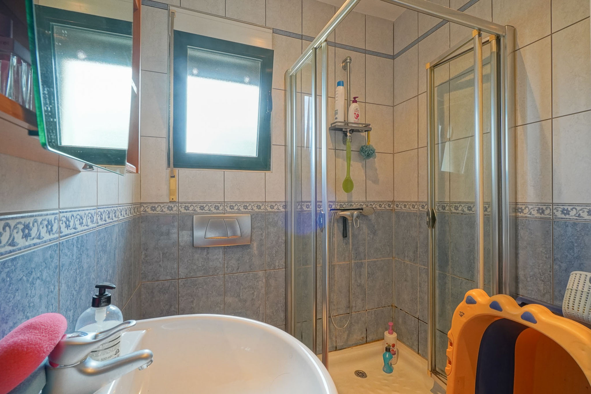 Praktische badkamer met keramische wastafel, glazen douchecabine en blauw-beige tegels. Een raam zorgt voor natuurlijke lichtinval.