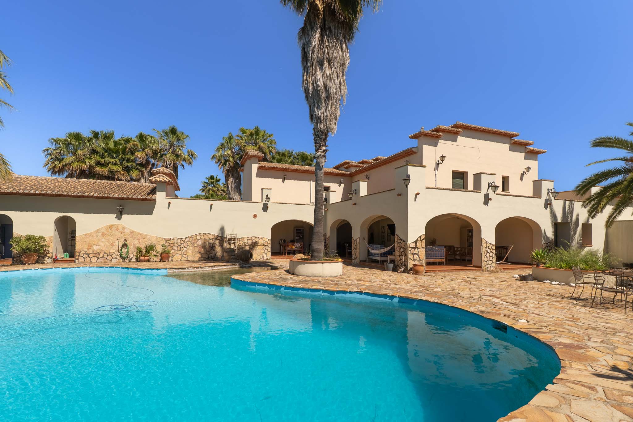 Ruim privézwembad met onregelmatige vorm en helderblauw water, omgeven door een uitgestrekt terras van natuursteen. De mediterrane villa heeft bogen en pannendaken, te midden van palmbomen.