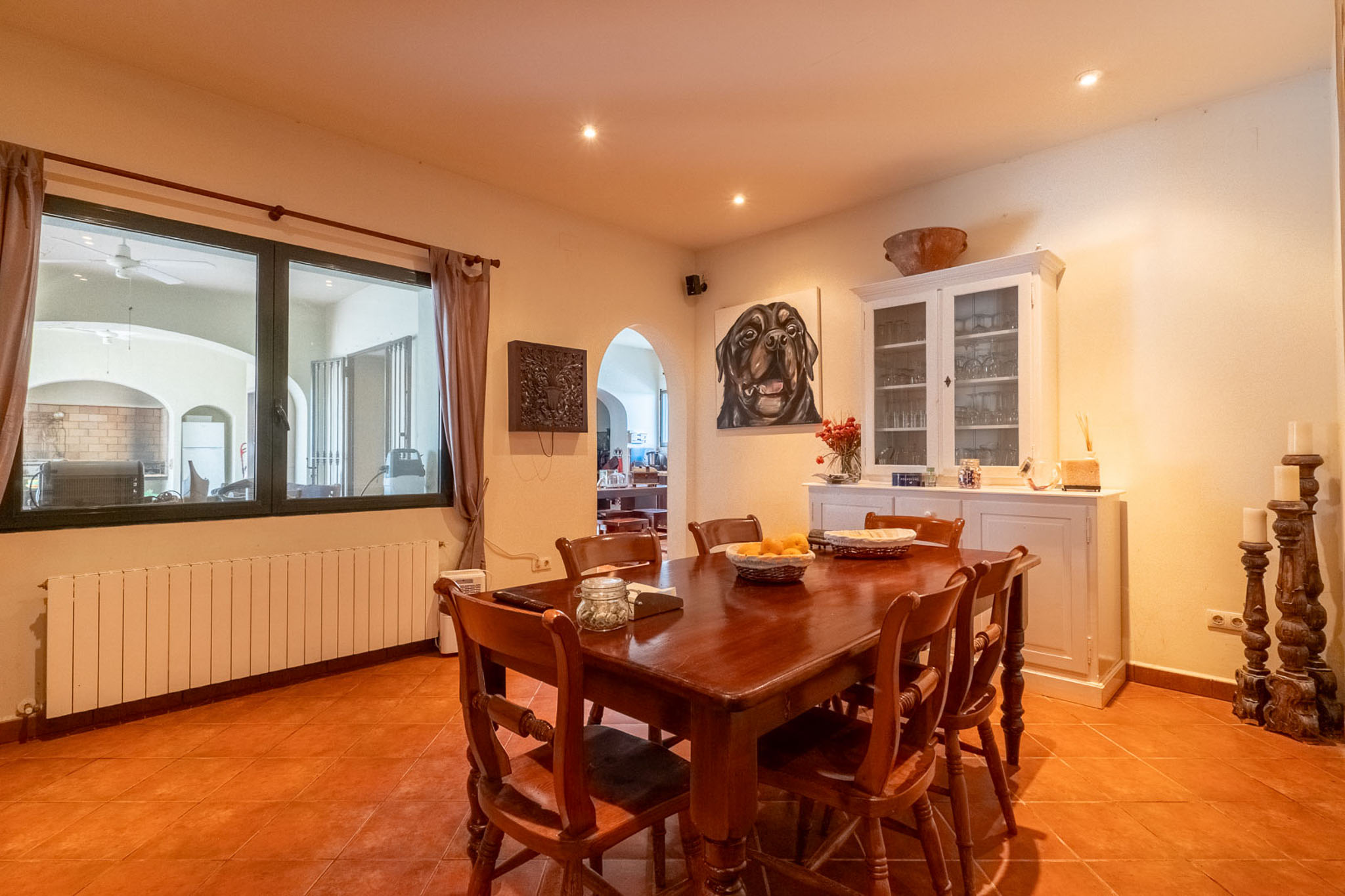Ruime eetkamer met een donkere houten tafel en bijpassende stoelen, op terracottategels. De ruimte heeft een radiator onder het raam en een witte vitrinekast.