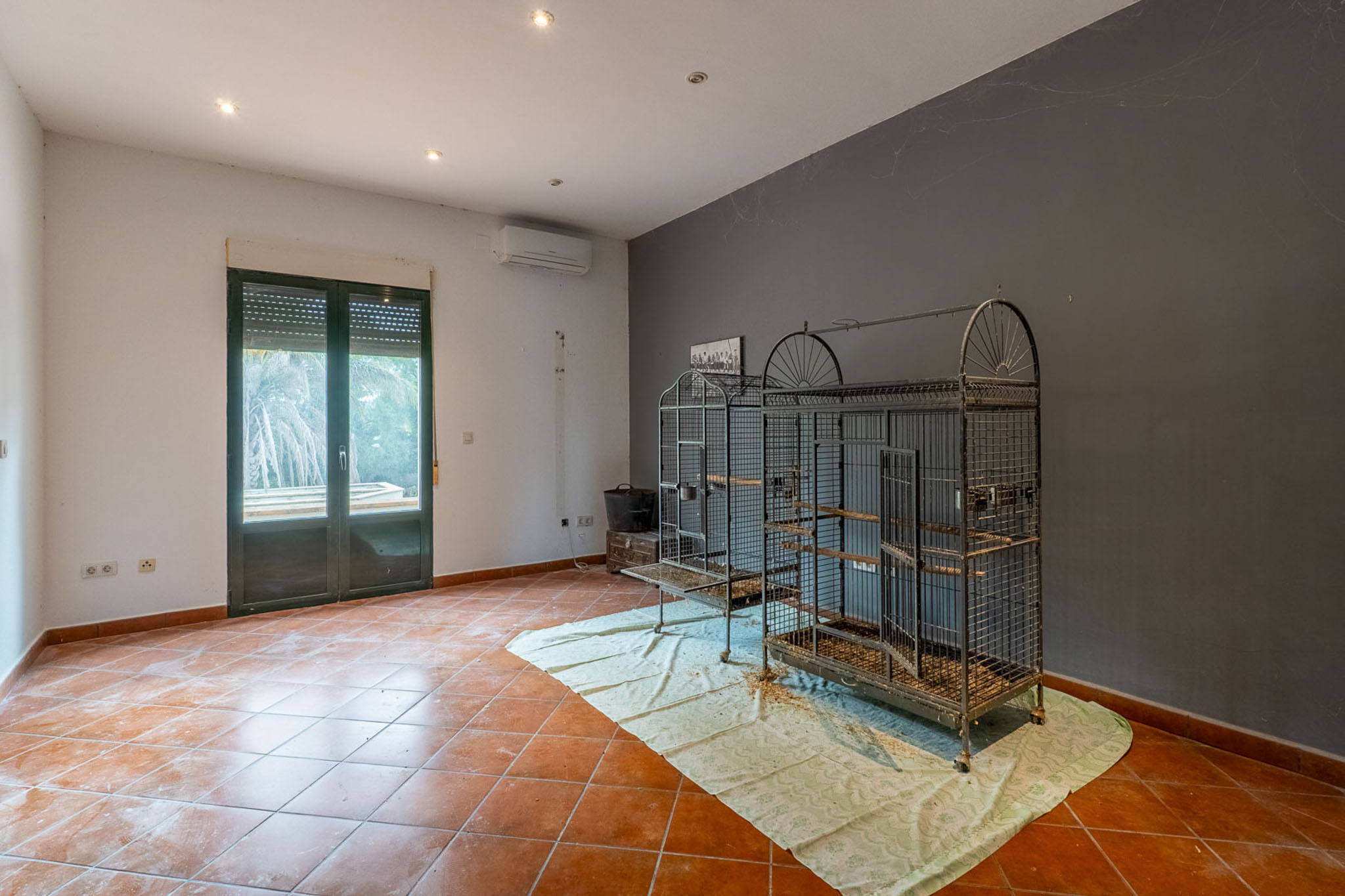 Ruime kamer met terracotta tegels, tweekleurige muren, inbouwverlichting en airconditioning. Dubbele beglazing deur met rolluik en grote metalen vogelkooien zijn zichtbaar.