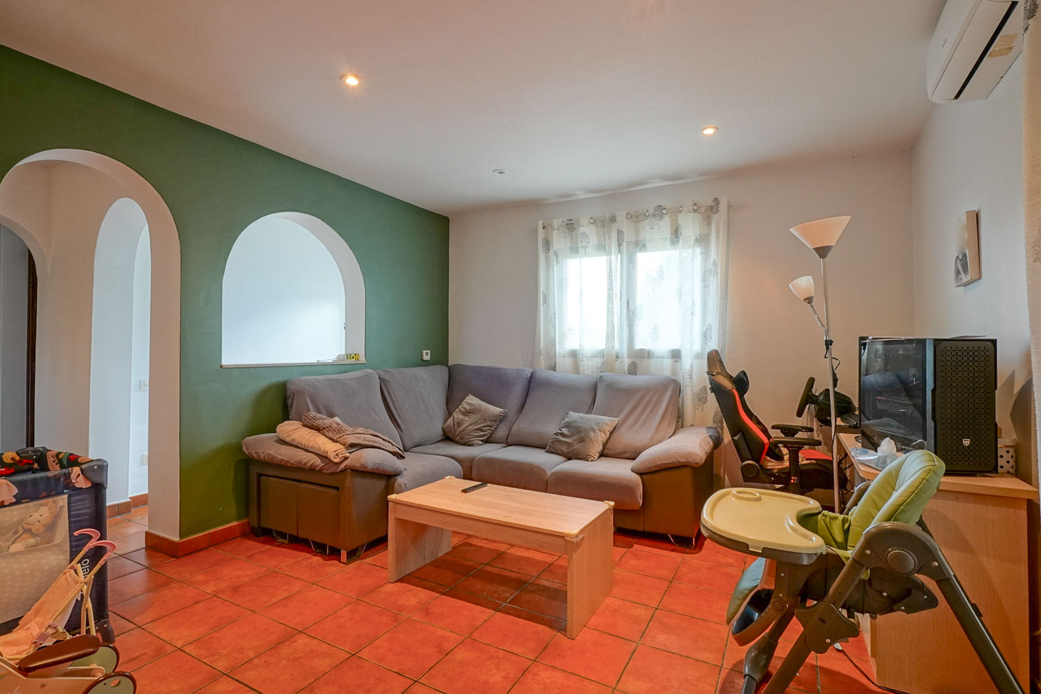 Ruime woonkamer met groene accentmuur en boogopeningen, terracotta tegelvloer en een comfortabele hoekbank. Functionele indeling met natuurlijk lichtinval.