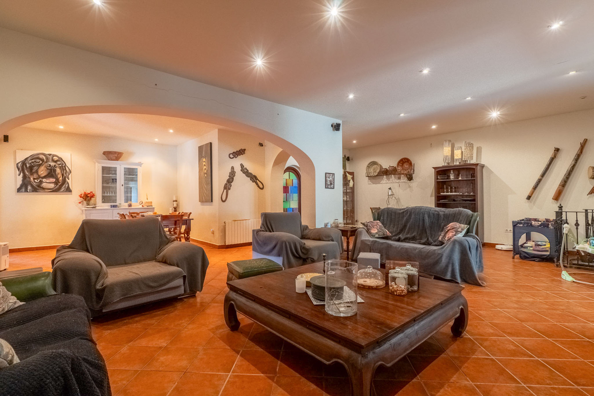 Ruime woonkamer met terracotta tegelvloer en inbouwspots in het plafond. Voorzien van een grote bank, fauteuils en een gesneden houten salontafel, verbonden met een eetruimte via bogen.