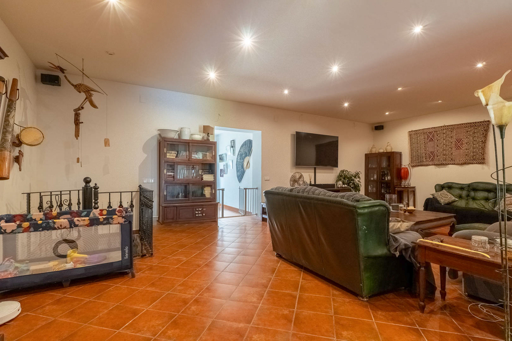 Ruime woonkamer met terracotta tegelvloer, inbouwspots, donkere leren banken en houten vitrinekasten. Een wand-tv en open doorgang naar de trap zijn zichtbaar.