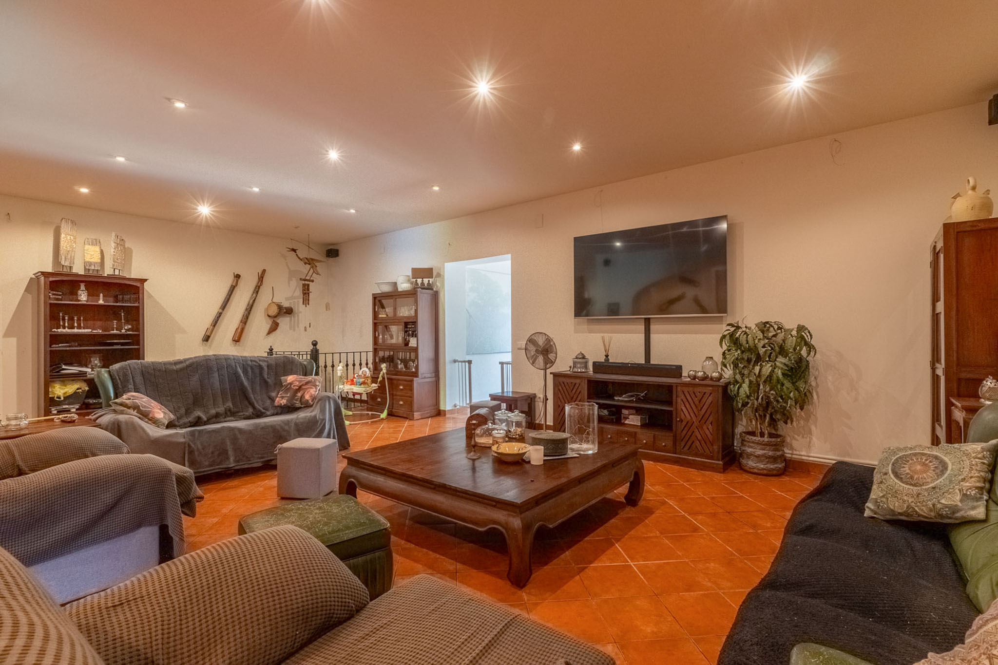 Ruime woonkamer met terracotta vloertegels, inbouwspots in het plafond en een grote wand-tv. Ingericht met donkerhouten meubels en bedekte banken, wat een warme sfeer creëert.