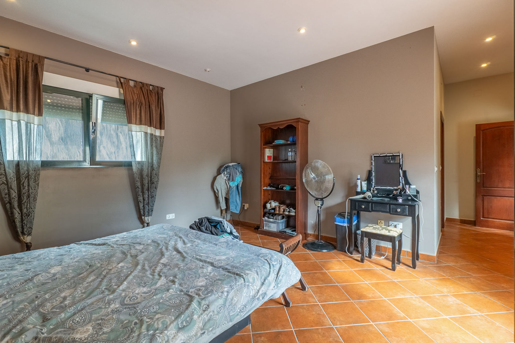Slaapkamer met bed, ramen met bruine gordijnen en rolluiken, en terracotta tegelvloer. Voorzien van een zwarte kaptafel, houten boekenkast en staande ventilator.
