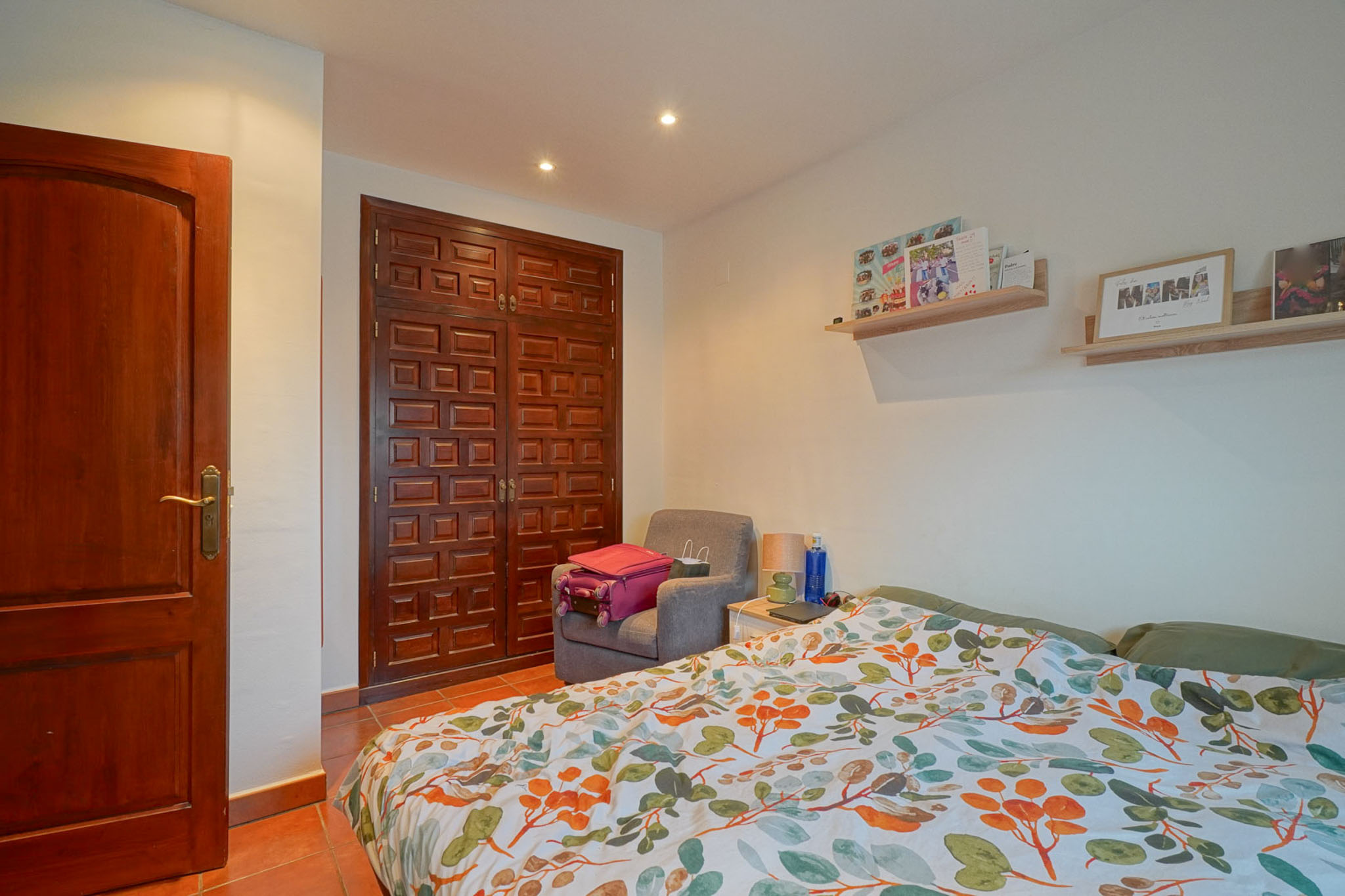 Slaapkamer met donkere paneelhouten kledingkast en deur, terracotta vloertegels en bed met levendig bladpatroon dekbed. Inbouwverlichting in het plafond.