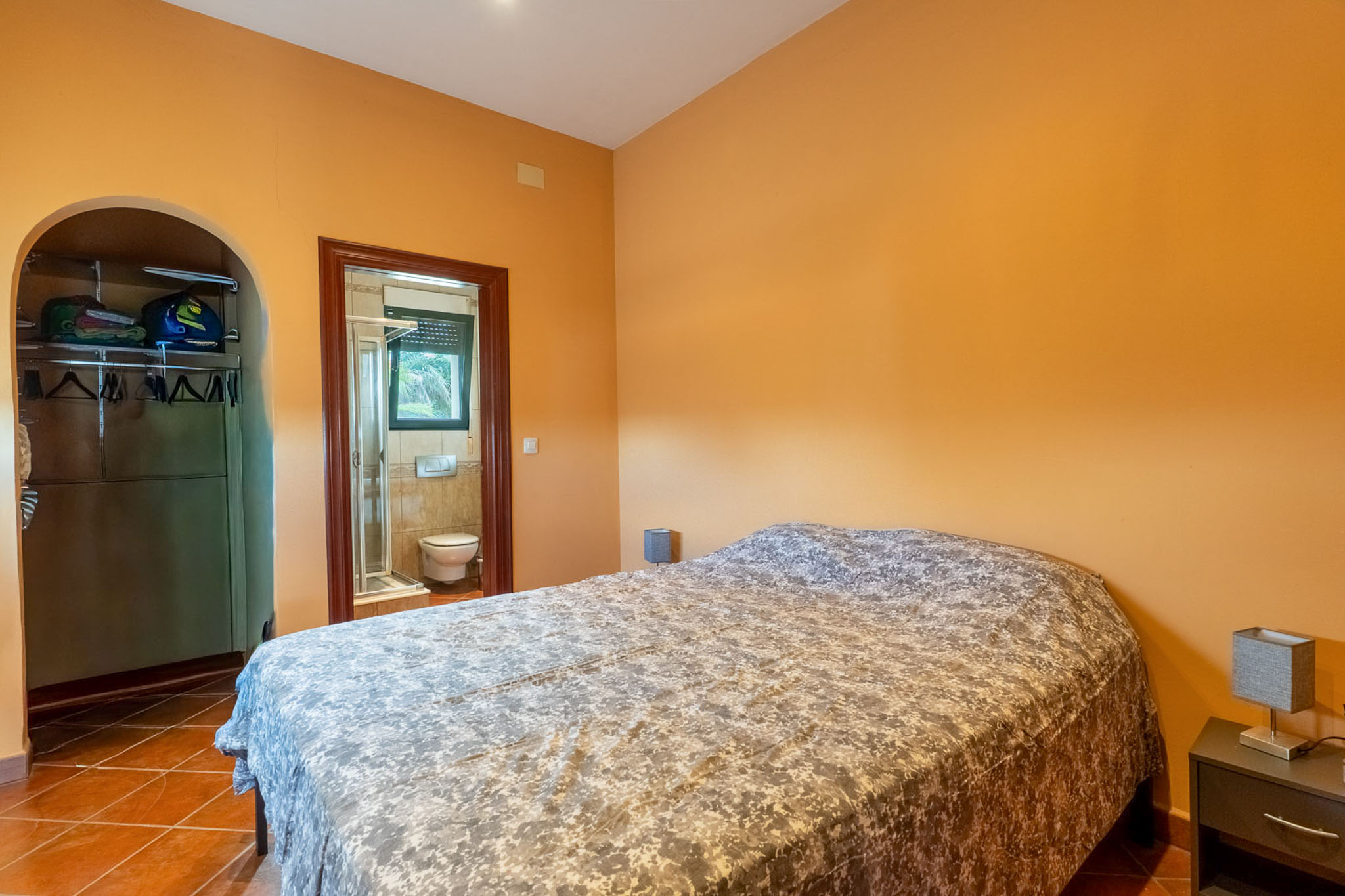 Slaapkamer met warme oranje muren en terracotta tegelvloer, voorzien van een tweepersoonsbed. Een open kledingkast onder een boog en een en-suite badkamer zijn zichtbaar.