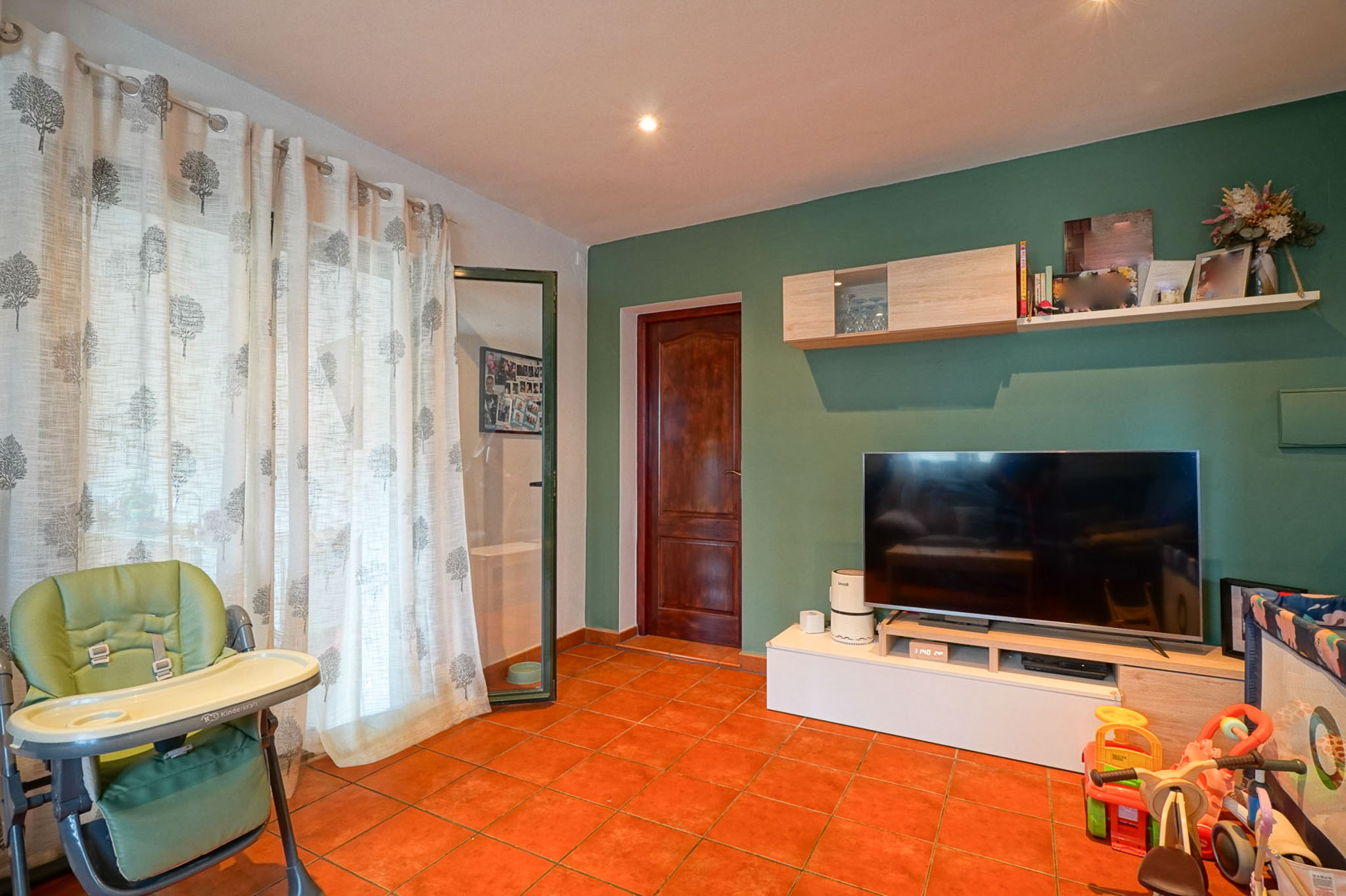 Woonruimte met terracottategels, een groene accentmuur en een tv op een modern meubel met zwevende planken. Een groot raam is voorzien van witte gordijnen met boompatroon.