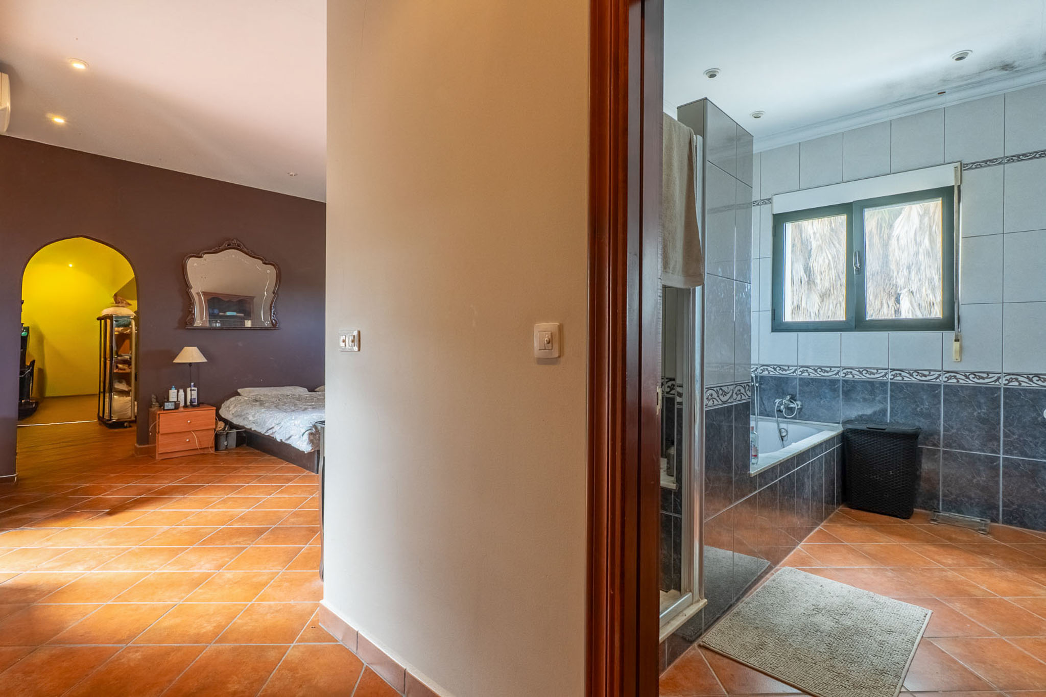 Zicht vanuit de gang met terracotta tegels, links een slaapkamer met donkere muren en sierlijke spiegel, rechts een betegelde badkamer met badkuip.