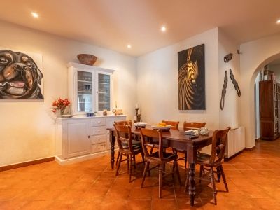 Eetkamer met donkere houten tafel en zes stoelen, terracotta tegelvloer en witte muren met inbouwspots. Een witte vitrinekast en twee decoratieve schilderijen sieren de ruimte.