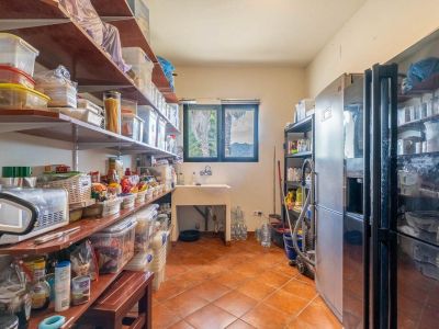 Functionele bijkeuken met uitgebreide schappen, een spoelbak en twee grote koelkasten. Terracotta tegelvloer en een raam met uitzicht op palmbomen en bergen.