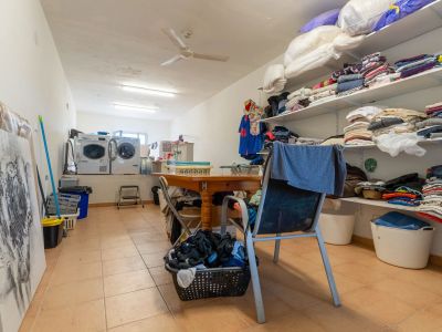 Functionele wasruimte met twee wasmachines, droger en open planken voor wasgoed. Een centrale houten tafel biedt een praktische werkruimte, aangevuld met tegelvloer en plafondventilator.