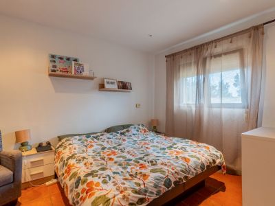 Lichte slaapkamer met tweepersoonsbed, kleurrijk bladpatroon dekbed en terracotta tegelvloer. Natuurlijk licht valt binnen via een raam met vitrage, aangevuld met een nachtkastje en wandplanken.