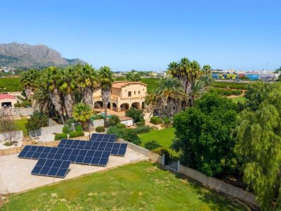 Luchtfoto van de traditionele villa, omringd door palmbomen en weelderige tuinen, met een opvallende zonnepaneleninstallatie en verre berguitzichten onder een heldere hemel.
