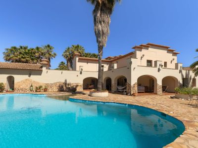 Ruim privézwembad met onregelmatige vorm en helderblauw water, omgeven door een uitgestrekt terras van natuursteen. De mediterrane villa heeft bogen en pannendaken, te midden van palmbomen.