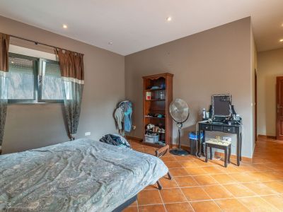 Slaapkamer met bed, ramen met bruine gordijnen en rolluiken, en terracotta tegelvloer. Voorzien van een zwarte kaptafel, houten boekenkast en staande ventilator.
