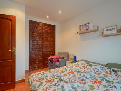 Slaapkamer met donkere paneelhouten kledingkast en deur, terracotta vloertegels en bed met levendig bladpatroon dekbed. Inbouwverlichting in het plafond.