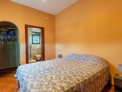 Slaapkamer met warme oranje muren en terracotta tegelvloer, voorzien van een tweepersoonsbed. Een open kledingkast onder een boog en een en-suite badkamer zijn zichtbaar.