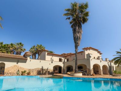 Uitgestrekt privézwembad met helder water, geflankeerd door een mediterrane villa met bogen en hoge palmbomen onder een blauwe lucht.