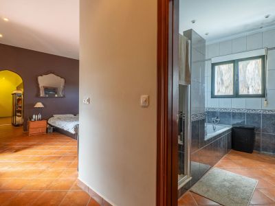 Zicht vanuit de gang met terracotta tegels, links een slaapkamer met donkere muren en sierlijke spiegel, rechts een betegelde badkamer met badkuip.