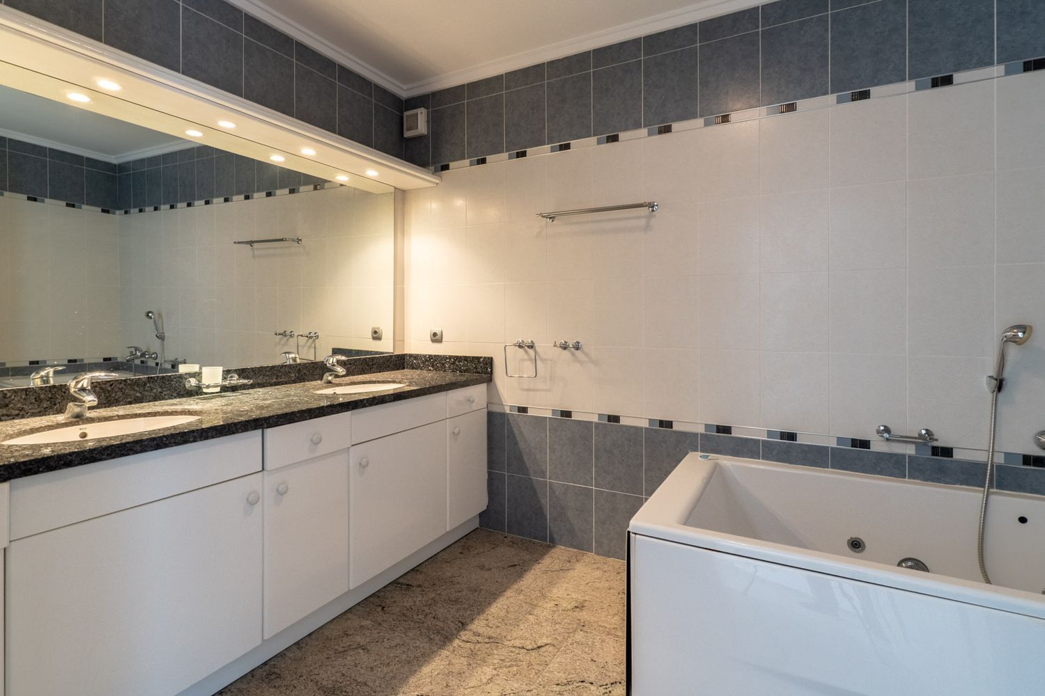 Badkamer met dubbele wastafel, donker granieten blad en witte kasten. Voorzien van een inbouwbad en een grote verlichte spiegel, met grijs-witte tegels.