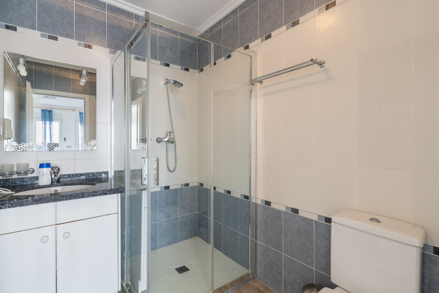 Badkamer met een donker gespikkelde granieten wastafelblad, een decoratieve spiegel en een glazen douchecabine. Muren zijn betegeld met lichtbeige en blauwgrijze tegels met een mozaïekrand.