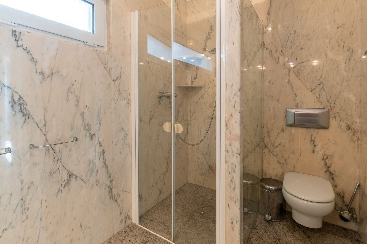 Badkamer met geaderde marmeren wanden en granieten vloer. Voorzien van een glazen douchecabine met geïntegreerde plank en een wandcloset, verlicht door een hooggeplaatst raam.