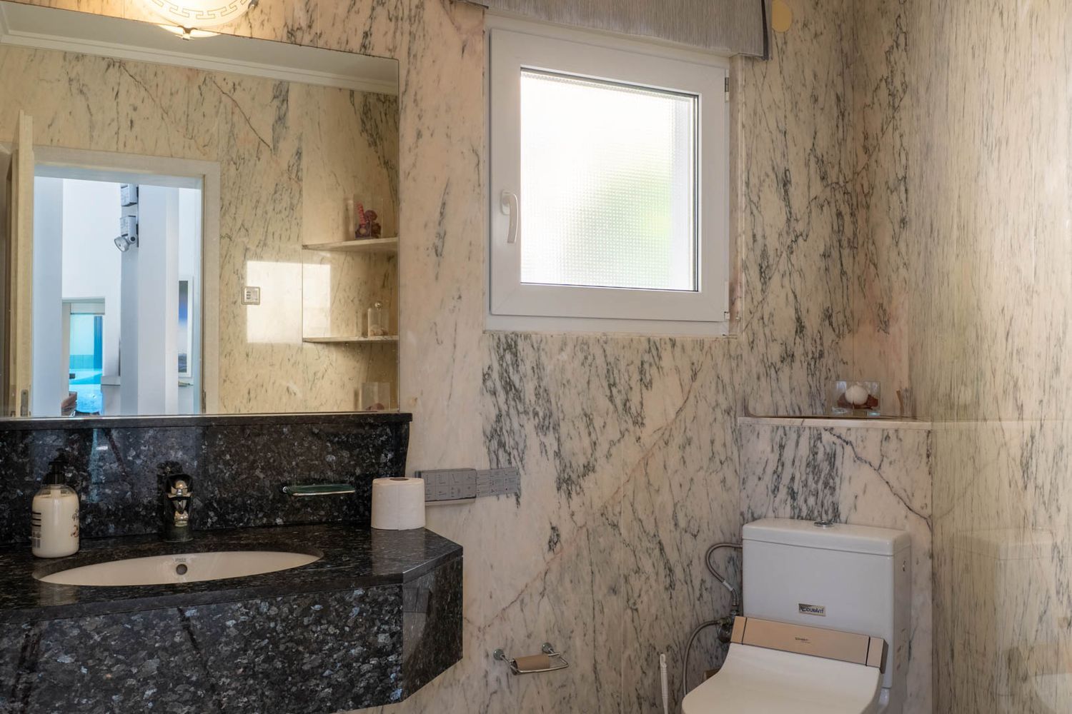Badkamer met licht geaderde marmeren muren, een donker granieten wastafelmeubel met geïntegreerde wastafel en een grote spiegel. Voorzien van een matglazen raam en toilet met bidetfunctie.