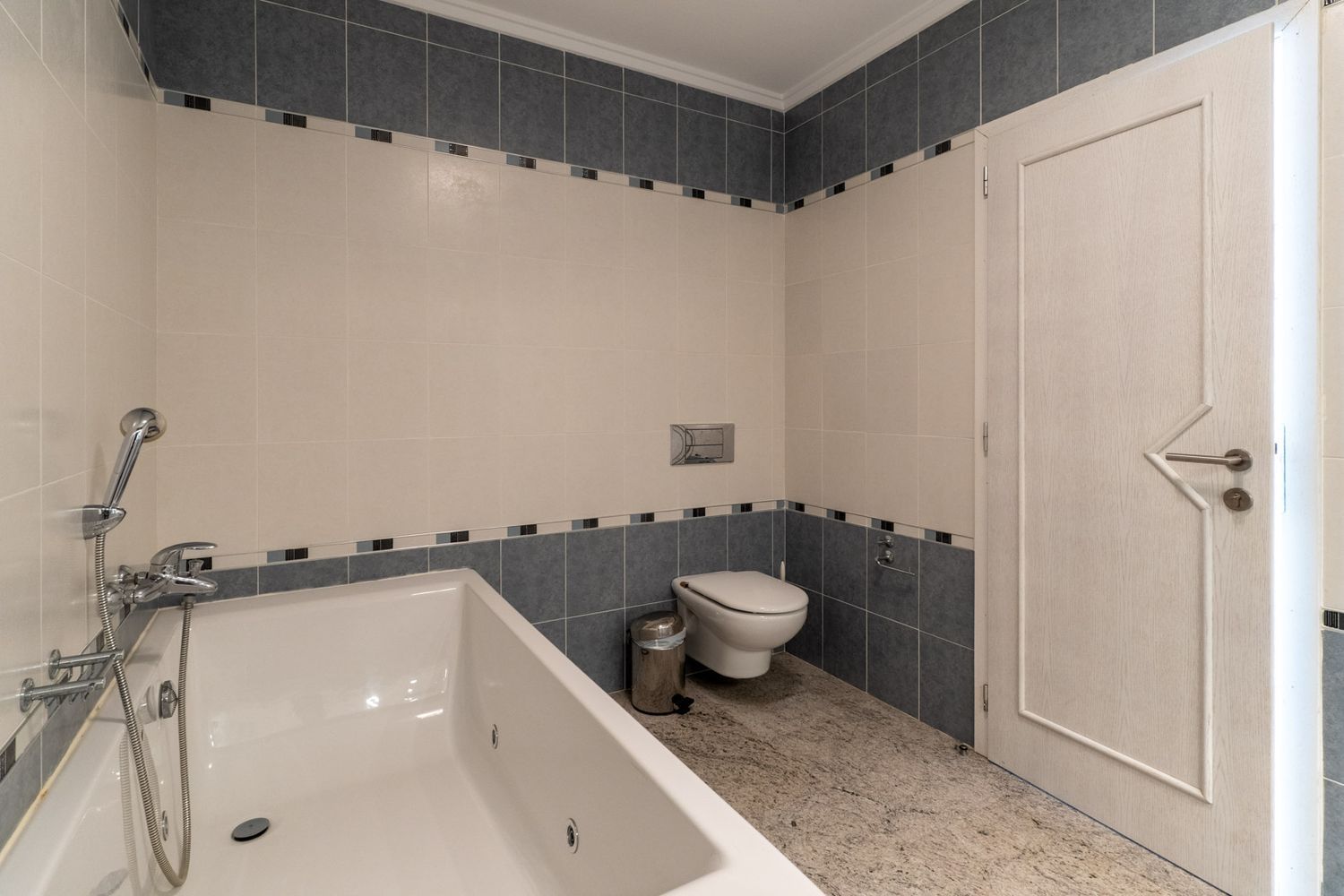 Badkamer met wit ligbad en geïntegreerde douche, wanden betegeld in beige en grijstinten met decoratieve sierrand. Lichte granieten vloer, zwevend toilet en een deur met geometrisch patroon.