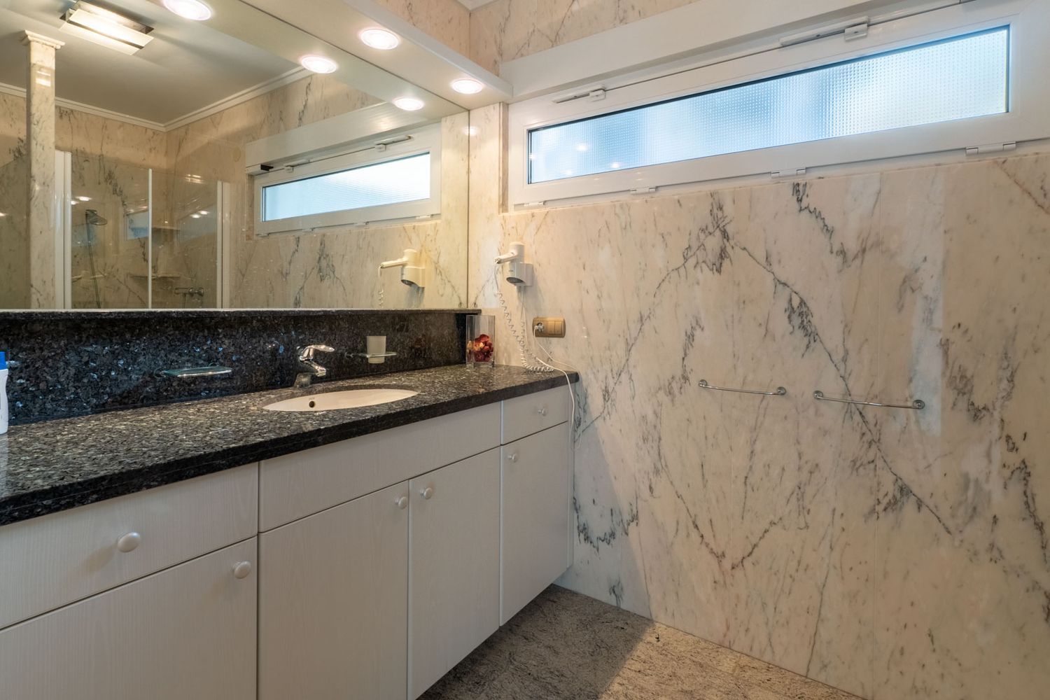 Elegante badkamer met geaderde marmeren wanden en vloer, en een wastafelmeubel met donker granieten blad. Een horizontaal matglazen raam biedt natuurlijk licht en privacy.