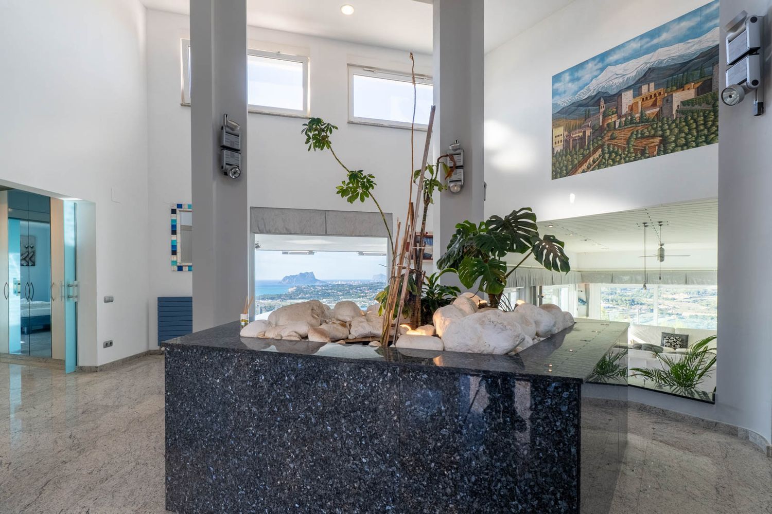 Entreehal met een decoratief eiland van donker graniet, witte stenen en planten. Hoge plafonds en grote ramen bieden panoramisch uitzicht op zee en de Peñón de Ifach.