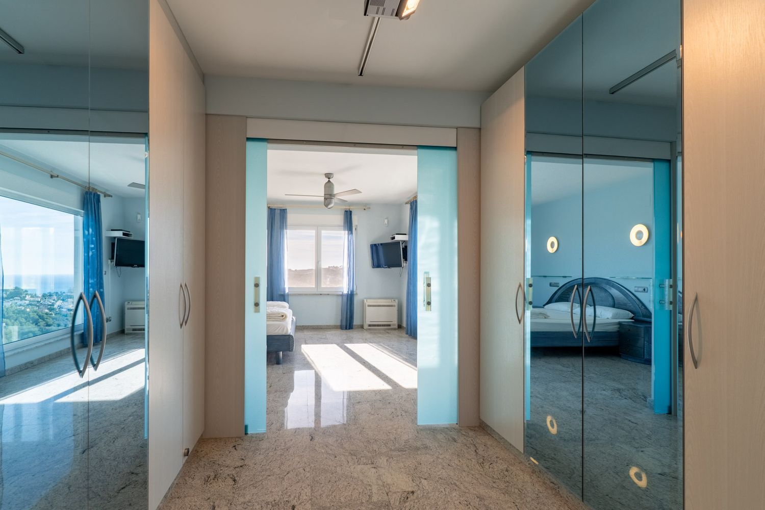 Lichte gang met granieten vloer en lichte houten kasten. Glazen schuifdeuren omlijsten een slaapkamer met zeezicht, terwijl spiegelpanelen een andere blauwgetinte slaapkamer weerspiegelen.