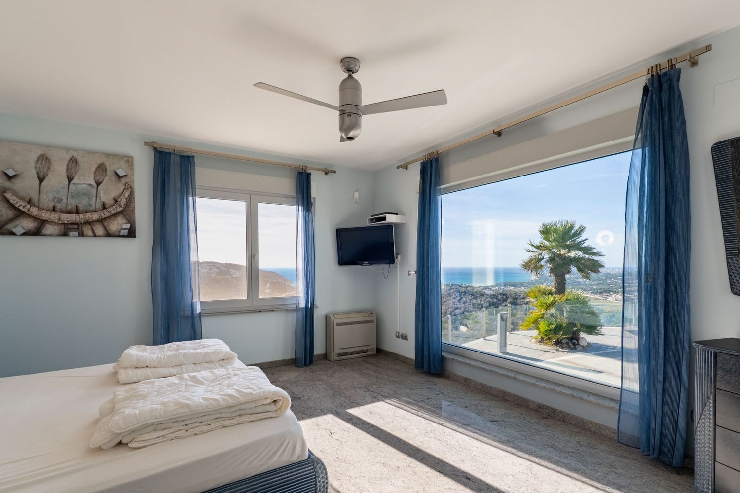 Lichte slaapkamer met groot panoramisch raam, direct zeezicht en een palmboom. Lichte graniet-effect tegelvloer en blauwe vitrage.