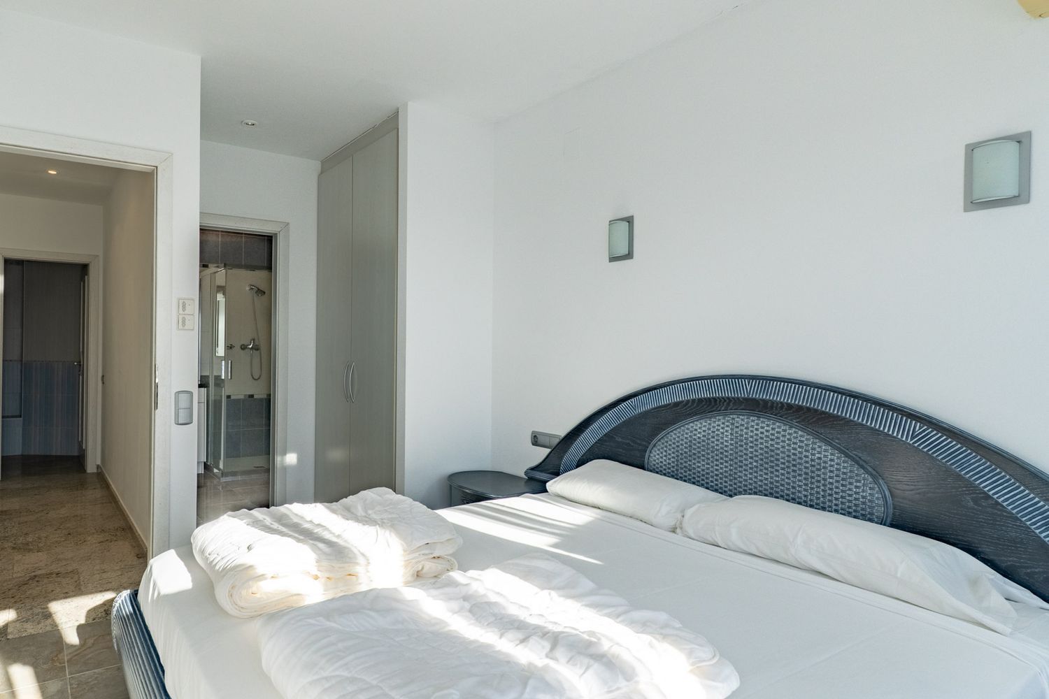 Lichte slaapkamer met tweepersoonsbed en donkerhouten hoofdbord met gevlochten inleg. Voorzien van een inbouwkast en directe toegang tot een badkamer met glazen douche.
