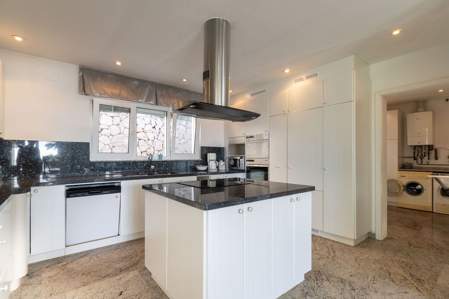 Moderne keuken met witte kasten, donkere granieten werkbladen en kookeiland met geïntegreerde kookplaat. Inbouwapparatuur, uitzicht op stenen muur vanuit raam, directe toegang tot wasruimte.