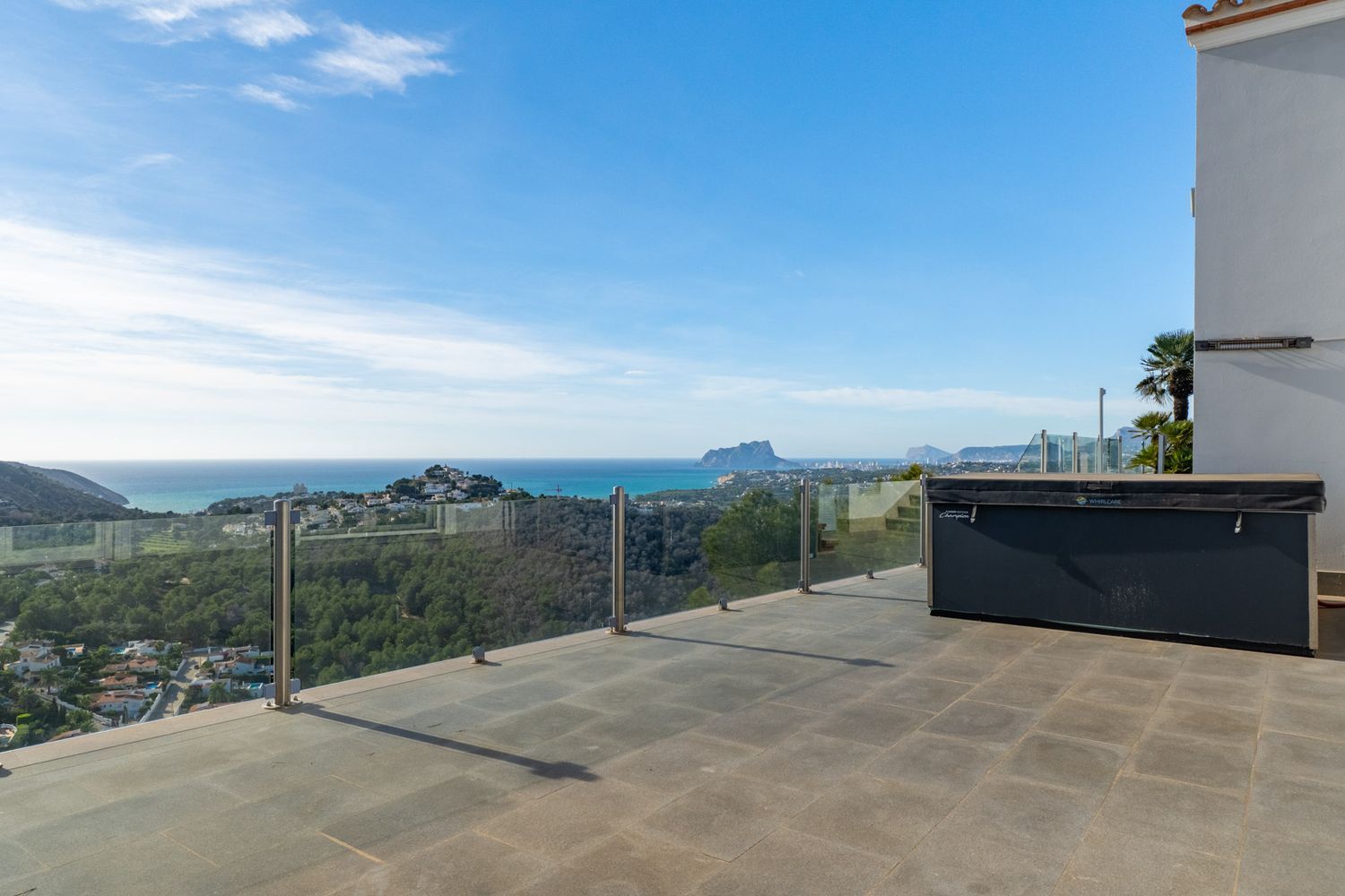 Ruim buitenterras met grijze tegels en een overdekte jacuzzi, omkaderd door een glazen balustrade met panoramisch uitzicht op zee en de Peñón de Ifach.
