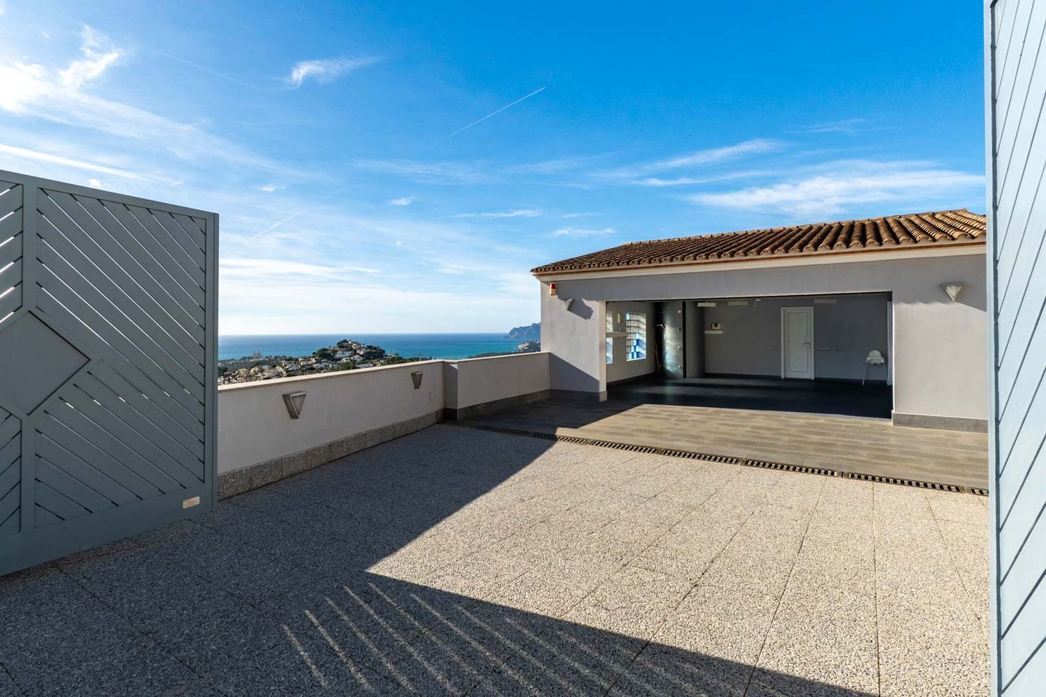 Ruime buitenruimte met tegelbestrating en toegang tot een overdekte garage. Biedt panoramisch uitzicht op de Middellandse Zee en de kust, omkaderd door een modern grijs hek en een lage muur met verlichting.